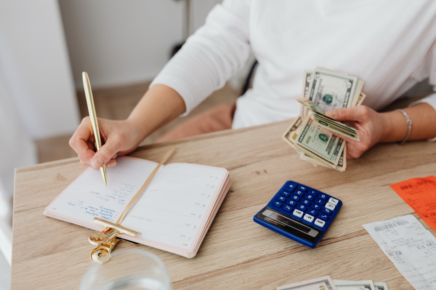 A person counts cash and writes in a notebook beside a calculator, tracking household finances