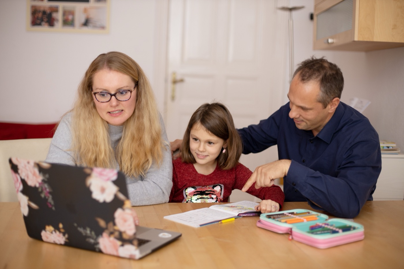 Two parents sit with their young daughter at a kitchen table, helping her with homework on a laptop